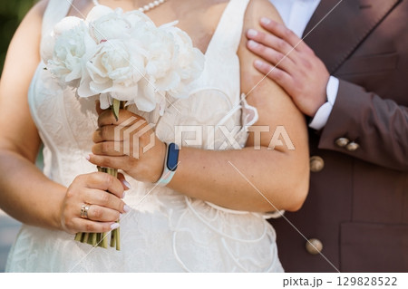 Intimate Wedding Embrace: Bride and Groom Holding Bouquet, White Peonies, Pearl Necklace, Brown Suit, Romantic Celebration, Close-Up View, Natural Lighting 129828522