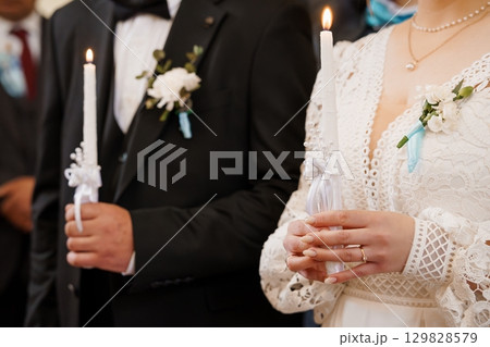 Orthodox Wedding Ceremony with Lit Candles: A Romantic Close-Up of Bride and Groom's Hands Holding Candles, White Lace, Traditional Attire Orthodox Wedding Ceremony with Lit Candles: A Romantic Close-Up of Bride and Groom's Hands Holding Candles, White Lace, Traditional Attire 129828579