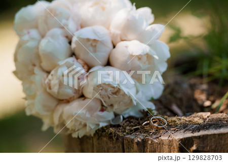 Wedding Rings on Wooden Log with White Peony Bouquet, Romantic Scene, Love, Golden Bands, Floral Arrangement, Soft Focus, Natural Light. 129828703