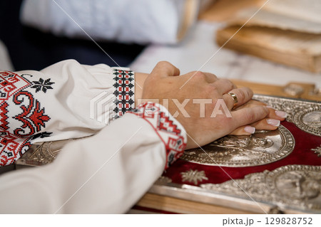 Hands Praying on Holy Book with Embroidered Sleeves, Orthodox Christian Tradition, Golden Cover, Red Textile, Spiritual Devotion, Close-Up Hands Praying on Holy Book with Embroidered Sleeves, Orthodox Christian Tradition, Golden Cover, Red Textile, Spiritual Devotion, Close-Up 129828752