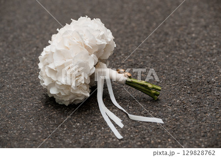 Wedding Bouquet and Rings on Asphalt: A Romantic Still Life with White Hydrangeas, Gold Bands, and Satin Ribbon against Dark Pavement. Wedding Bouquet and Rings on Asphalt: A Romantic Still Life with White Hydrangeas, Gold Bands, and Satin Ribbon against Dark Pavement. 129828762