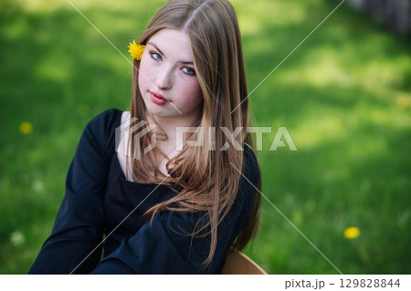 Teenage student girl sitting outdoors with a dandelion tucked behind her ear, relaxing on a grassy field during her school break 129828844