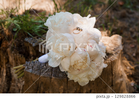 Romantic Wedding Rings Resting on White Peony Bouquet Placed on Tree Stump in Forest, Golden Bands, Soft Lighting, Love, Marriage, Engagement 129828866
