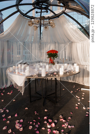 Romantic Dinner Setup in Glass Dome with Red Roses, Candles and Draped White Fabric. Intimate Evening Mood, Dark Wood Table, Warm Lighting. Romantic Dinner Setup in Glass Dome with Red Roses, Candles and Draped White Fabric. Intimate Evening Mood, Dark Wood Table, Warm Lighting. 129828872