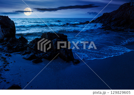 landscape by the sea at night in summer. beautiful beach with rocks washed by calm waves in full moon light. view to horizon under dark sky with clouds. scenic travel destination for leisure and relax 129829605