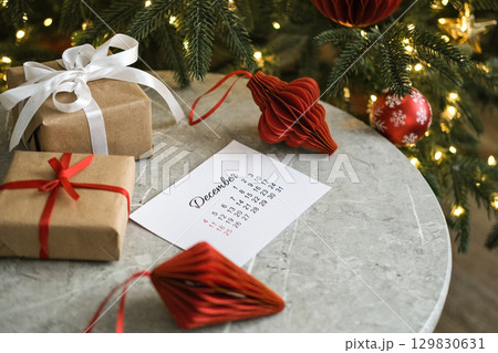 December calendar surrounded by Christmas gifts and red Christmas tree decorations on marble table near Christmas tree 129830631
