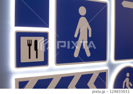 Various directional and informational signs. Symbols include pedestrian icon, dining symbol with utensils, and directional arrow, all set on blue background with illuminated edges Various directional and informational signs. Symbols include pedestrian icon, dining symbol with utensils, and directional arrow, all set on blue background with illuminated edges 129833031