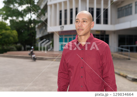 Confident bald Italian man in burgundy shirt standing in front of university building on sunny day Confident bald Italian man in burgundy shirt standing in front of university building on sunny day 129834664