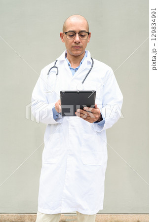 Hispanic Male Doctor in Lab Coat with Stethoscope on Plain Background Using Tablet Computer Hispanic Male Doctor in Lab Coat with Stethoscope on Plain Background Using Tablet Computer 129834991