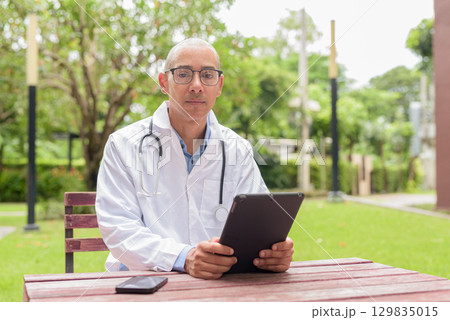 Doctor in uniform relaxing outdoors in hospital garden, smiling and confident using tablet computer 129835015
