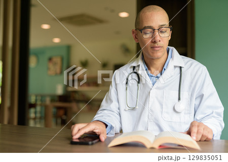 Doctor Sitting Indoors in Clinic or Hospital Area Library with Stethoscope Reading Book 129835051