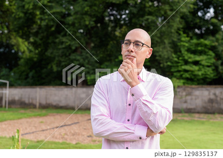 Bald Hispanic man thinking and looking up with hand on chin outdoors Bald Hispanic man thinking and looking up with hand on chin outdoors 129835137