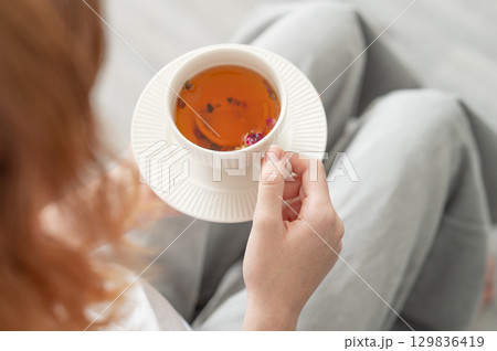 Woman holding white cup of tea with lemon and herbs in cozy bright room. 129836419
