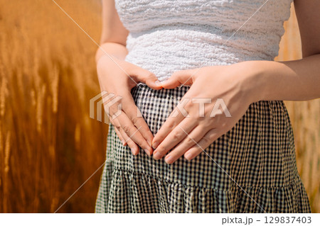 Pregnant Woman Making Heart Shape on Belly in Wheat Field 129837403