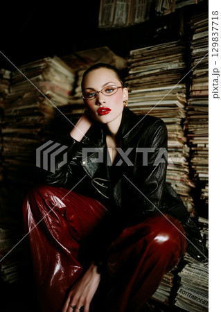 A woman in a black jacket and red pants sits on top of a pile of magazines 129837718