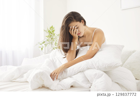 Unhappy young Caucasian woman sitting in bed, feeling unwell after night and unwilling to get up. 129837772