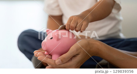 Saving Money. Child placing coin into piggy bank with parent guidance. 129839356