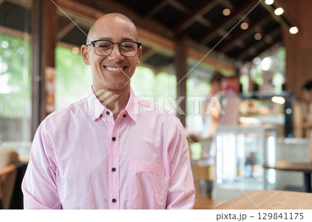 Happy bald Hispanic man wearing pink casual business shirt and eyeglasses in coffee shop cafe 129841175