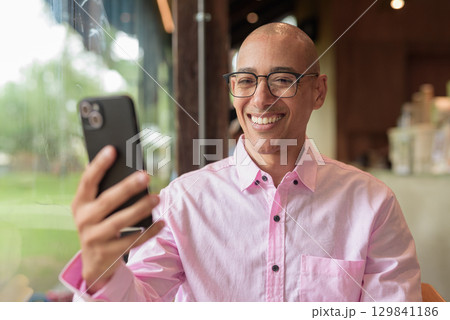 Bald Hispanic man wearing pink casual business shirt and eyeglasses in coffee shop using phone 129841186