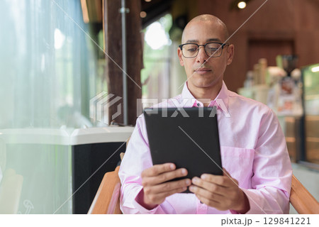 Bald mature Hispanic man wearing pink casual business shirt in coffee shop using tablet computer 129841221