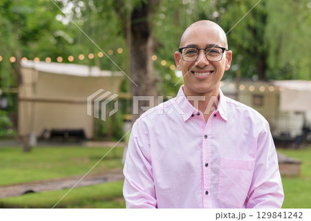 Happy bald Hispanic man wearing pink casual business shirt and eyeglasses outdoors smiling 129841242