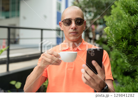 Bald Hispanic man face sitting outdoors at cafe restaurant wearing orange polo shirt using phone 129841282
