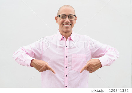 Bald Hispanic man wearing pink casual business shirt and eyeglasses pointing at self 129841382