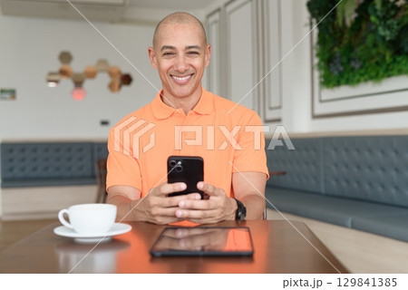 Bald Hispanic businessman having coffee break in cafe and using phone Bald Hispanic businessman having coffee break in cafe and using phone 129841385