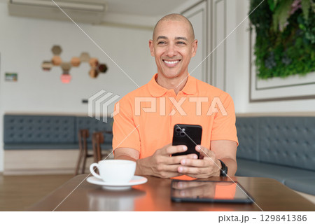 Bald Hispanic businessman having coffee break in cafe and using phone while thinking 129841386