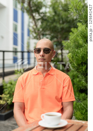 Bald Hispanic man face sitting outdoors at cafe restaurant wearing orange polo shirt 129841427