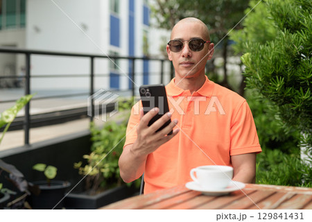 Bald Hispanic man face sitting outdoors at cafe restaurant wearing orange polo shirt using phone Bald Hispanic man face sitting outdoors at cafe restaurant wearing orange polo shirt using phone 129841431