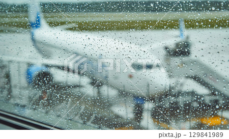 Raindrops blurring airport window, airplane resting on wet tarmac amid rainy weather conditions 129841989