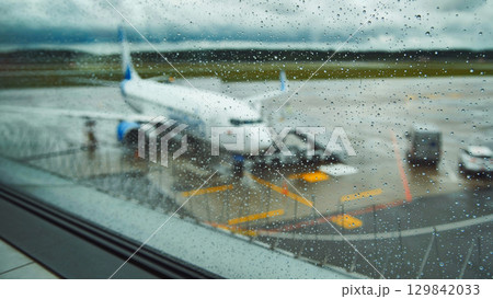 Raindrops blurring airport window view of parked airplane against gloomy runway backdrop Raindrops blurring airport window view of parked airplane against gloomy runway backdrop 129842033