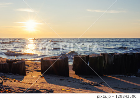 Wooden breakwater stretches into blue sea under soft evening light. Gentle waves touch sandy shore while sun reflects warmly on weathered timber posts and shimmering water 129842300