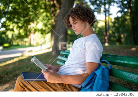 Curly-haired teenager sitting on the bench in the park and reading a book Curly-haired teenager sitting on the bench in the park and reading a book 129842306