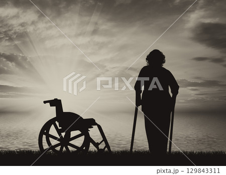 Silhouette of an old woman on crutches on a background of sea sunset and wheelchair 129843311