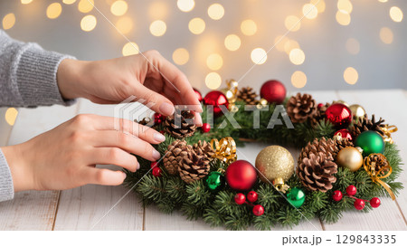 Hands decorating a Christmas wreath with pine cones, baubles, red berries, and gold ribbon on wooden table. Warm festive lights in background. Hands decorating a Christmas wreath with pine cones, baubles, red berries, and gold ribbon on wooden table. Warm festive lights in background. 129843335