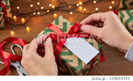 Christmas gift wrapping process with hands tying red ribbon on present with tag, festive lights glowing in soft background 129843343