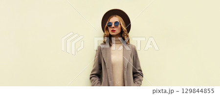 Beautiful stylish young woman, fashionable girl in round hat, brown jacket, glasses posing on wall 129844855