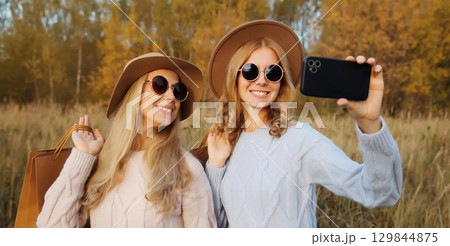 Happy women friends taking selfie with phone, shopping bags in autumn, girlfriends smiling in park 129844875