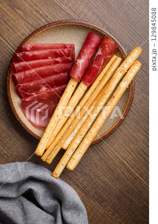 Slices of smoked bresaola and grissini sticks on plate on wooden table. Top view. Slices of smoked bresaola and grissini sticks on plate on wooden table. Top view. 129845088