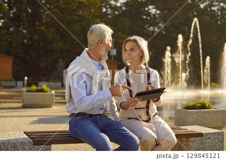 Senior couple sitting on bench in park using digital tablet enjoying retirement. Senior couple sitting on bench in park using digital tablet enjoying retirement. 129845121