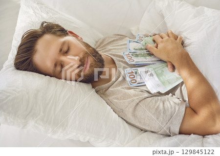 Smiling sleeping man lying in bed with closed eyes, embracing lots of cash dollar banknotes at home Smiling sleeping man lying in bed with closed eyes, embracing lots of cash dollar banknotes at home 129845122