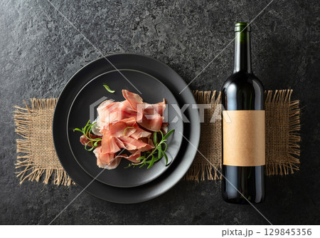 Bottle of red wine and prosciutto on a black plate. 129845356