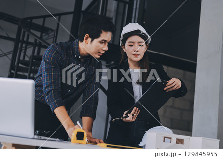 Construction manager and engineer dressed in orange work vests and hard helmets explore construction documentation on the building site near the steel frames 129845955