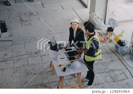 Construction manager and engineer dressed in orange work vests and hard helmets explore construction documentation on the building site near the steel frames 129845964