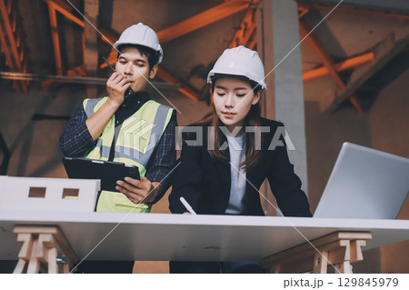 Construction manager and engineer dressed in orange work vests and hard helmets explore construction documentation on the building site near the steel frames Construction manager and engineer dressed in orange work vests and hard helmets explore construction documentation on the building site near the steel frames 129845979