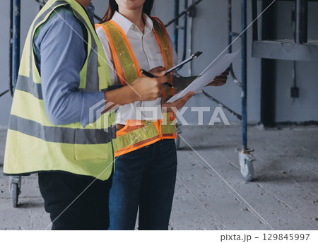Construction manager and engineer dressed in orange work vests and hard helmets explore construction documentation on the building site near the steel frames 129845997