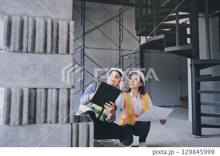 Construction manager and engineer dressed in orange work vests and hard helmets explore construction documentation on the building site near the steel frames Construction manager and engineer dressed in orange work vests and hard helmets explore construction documentation on the building site near the steel frames 129845999
