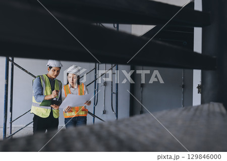 Construction manager and engineer dressed in orange work vests and hard helmets explore construction documentation on the building site near the steel frames 129846000